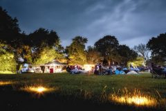 An early evening photo of a group of eventgoers relaxing on a field on camping chairs and blankets.