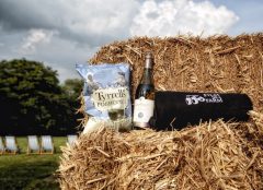 An arty photograph of a bottle of wine, Tyrrells Poshcorn and Film on a Farm blanket resting on a hay bale.