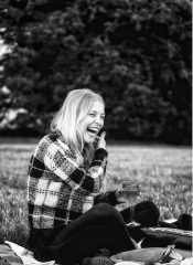 A young woman sat on a picnic blanket with a glass of wine laughing and smiling.