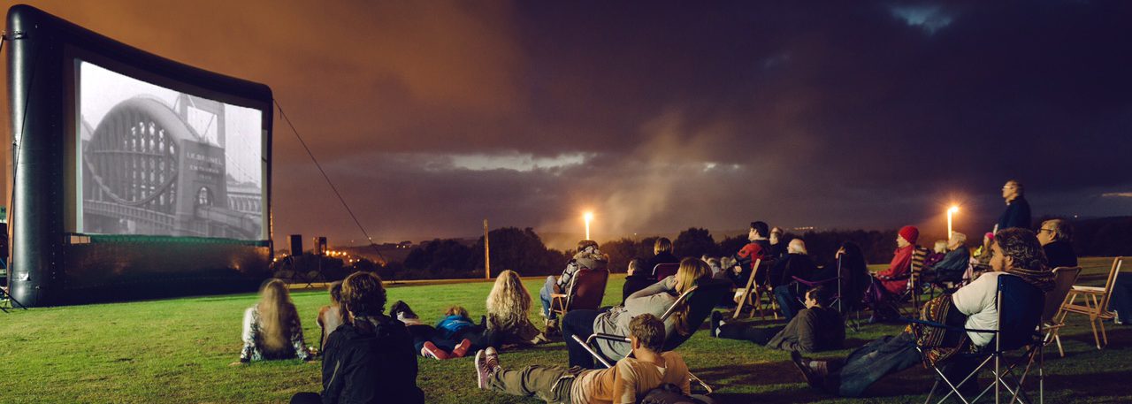 A group of revellers relax in a field to watch a film on a big screen.