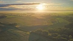 A photo of West Lodge Rural Centre, Kettering. The photo is an aerial shot, showing the scale of the countryside around the venue. The sun is setting in the background casting an orange glow across the rolling countryside.
