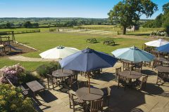 A photo showing the outside garden at Hilltop Farm Shop, Leamington Spa. In the foreground are a number of outside dining tables with chairs and umbrellas. In the background an enclosed field with a number of picnic benches and a children's play park.